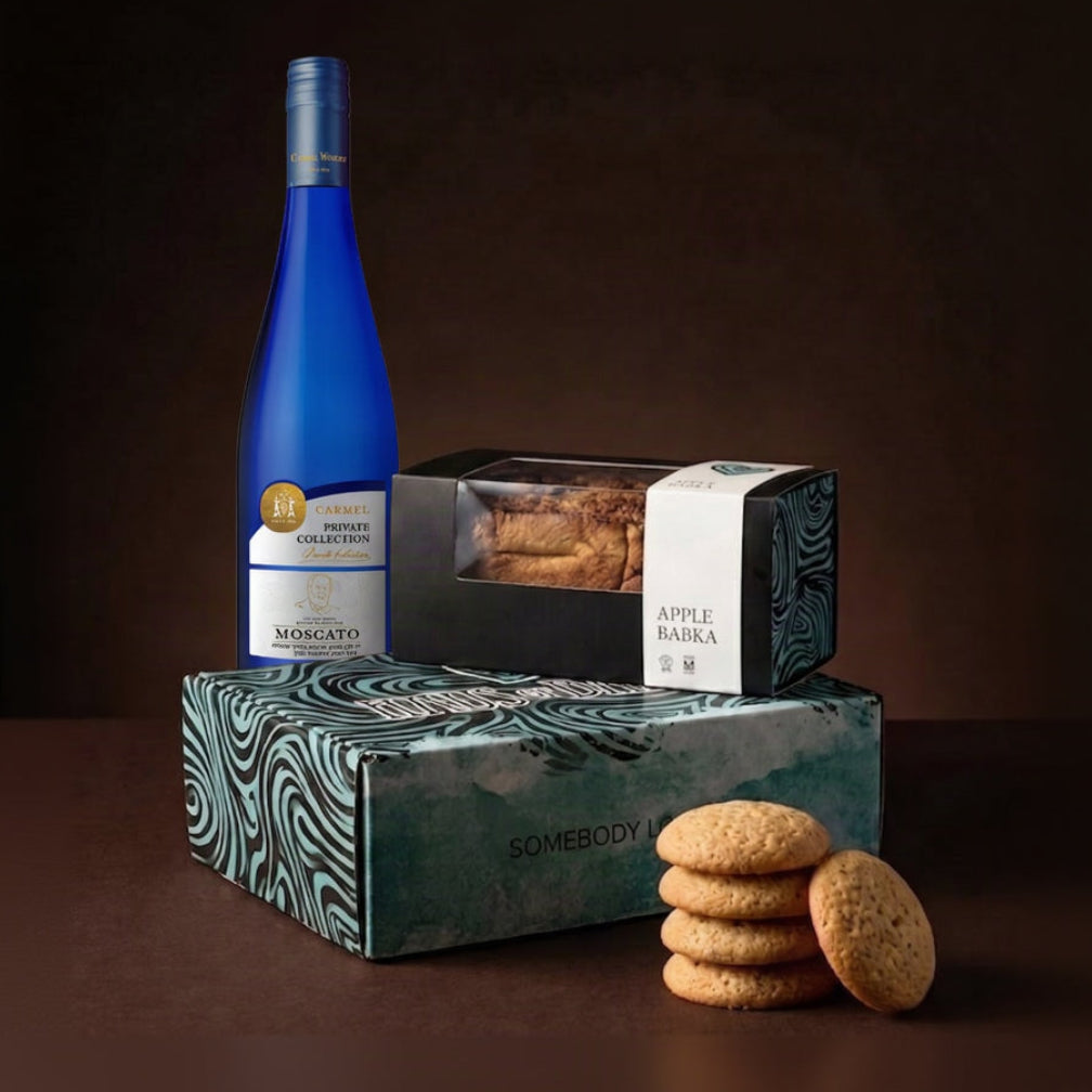 A curated gift set featuring a bottle of Moscato wine, Apple Babka cookies, and two decorated boxes, one blue and one teal with a wavy pattern. The set is styled with a stack of cookies on a dark brown background.