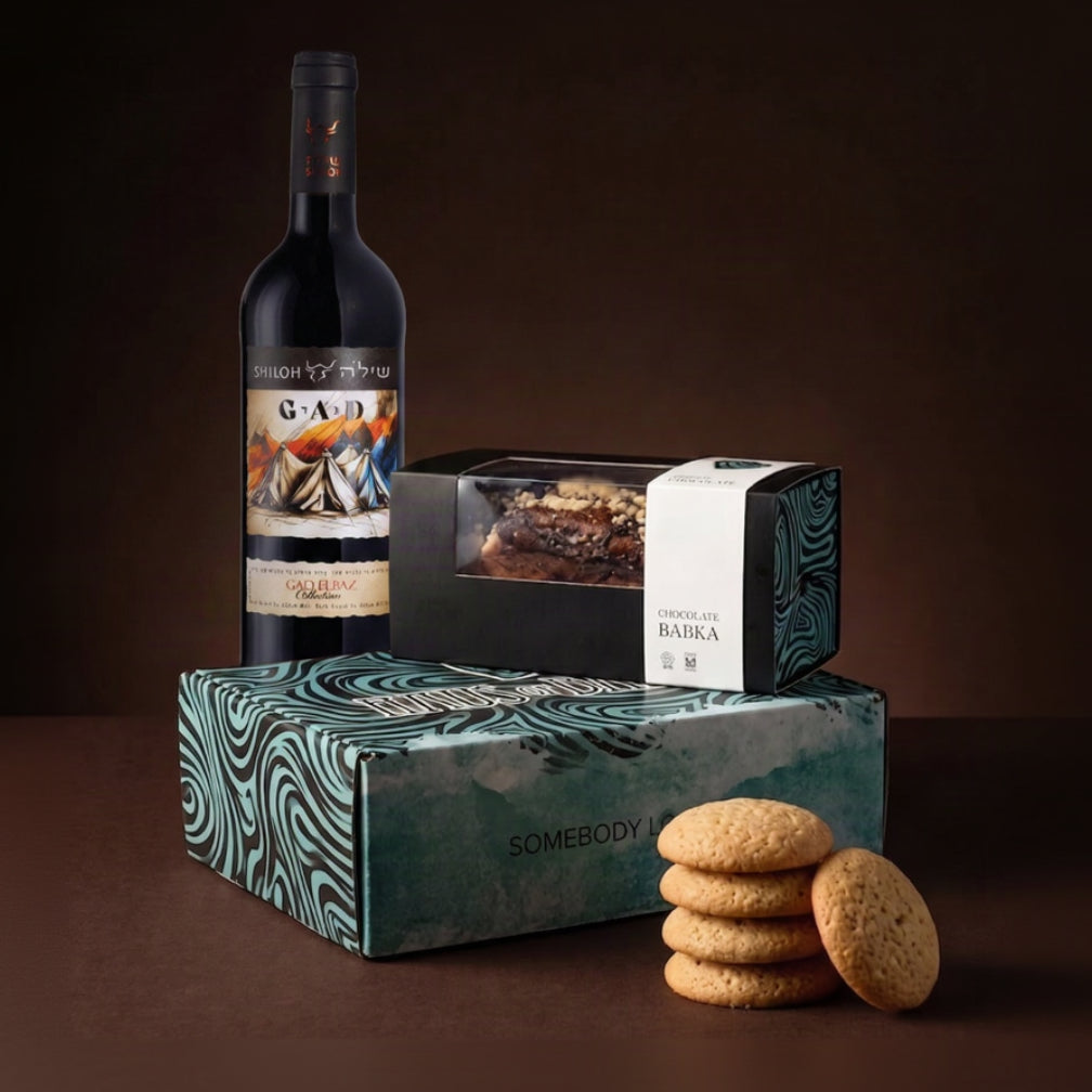 A curated gift set featuring a bottle of Shiloh Gad wine, a box of chocolate Babka, and two patterned gift boxes with cookies, arranged on a dark brown background.