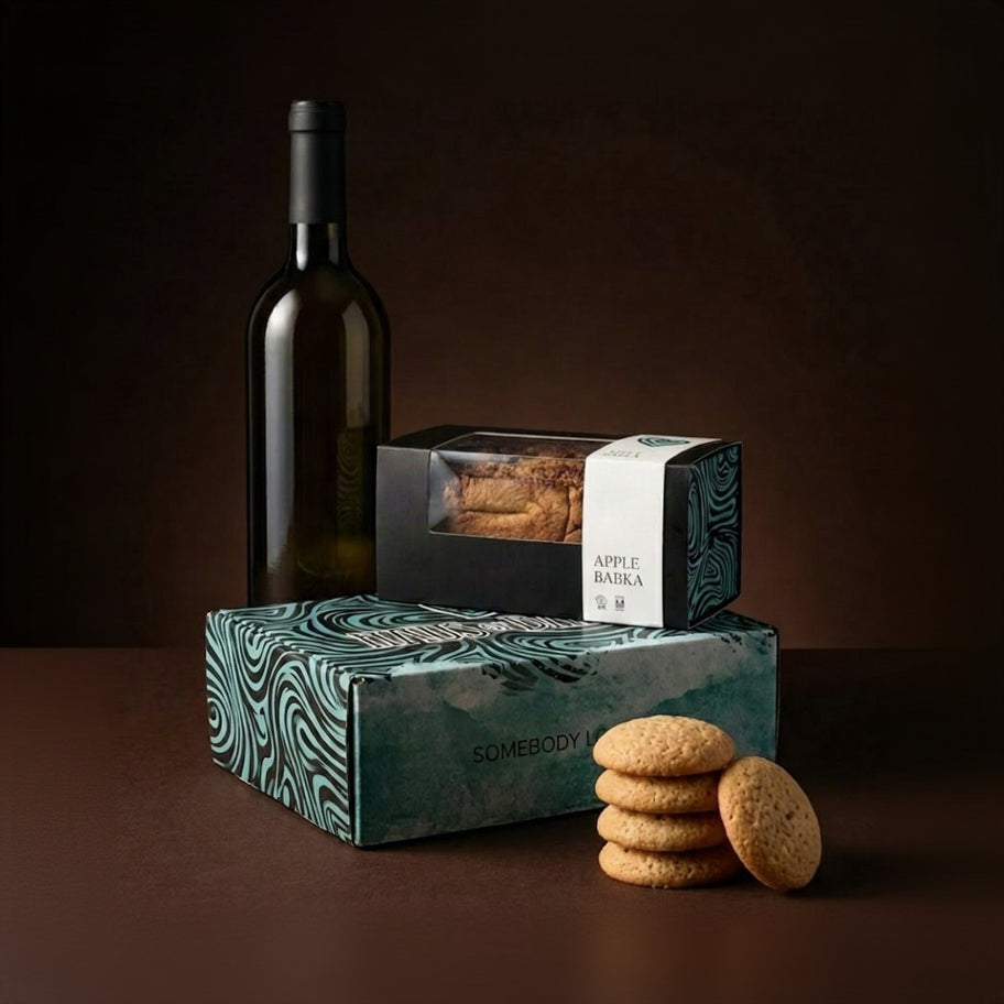 A gift set featuring a bottle of white wine, a box of Apple Bakka cookies, and a larger box of Sombody cookies with a stack of cookies in front, set against a dark brown background.