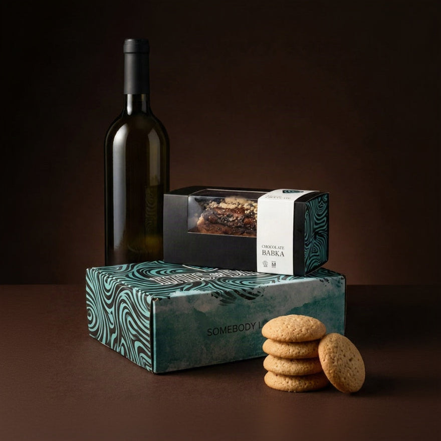 A gift set featuring a bottle of white wine, a chocolate bar in a window box, and cookies, all arranged with decorative teal and black patterned packaging on a dark brown background. The brand name 'SOMEBODY' is visible on the boxes.