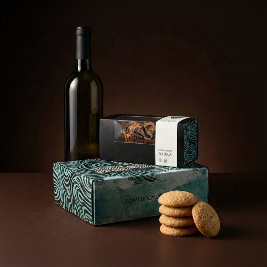 A gourmet gift set featuring a bottle of white wine, a box of chocolate-covered biscuits, and a larger box of chocolate cookies with three cookies displayed in front, all set against a dark brown background.