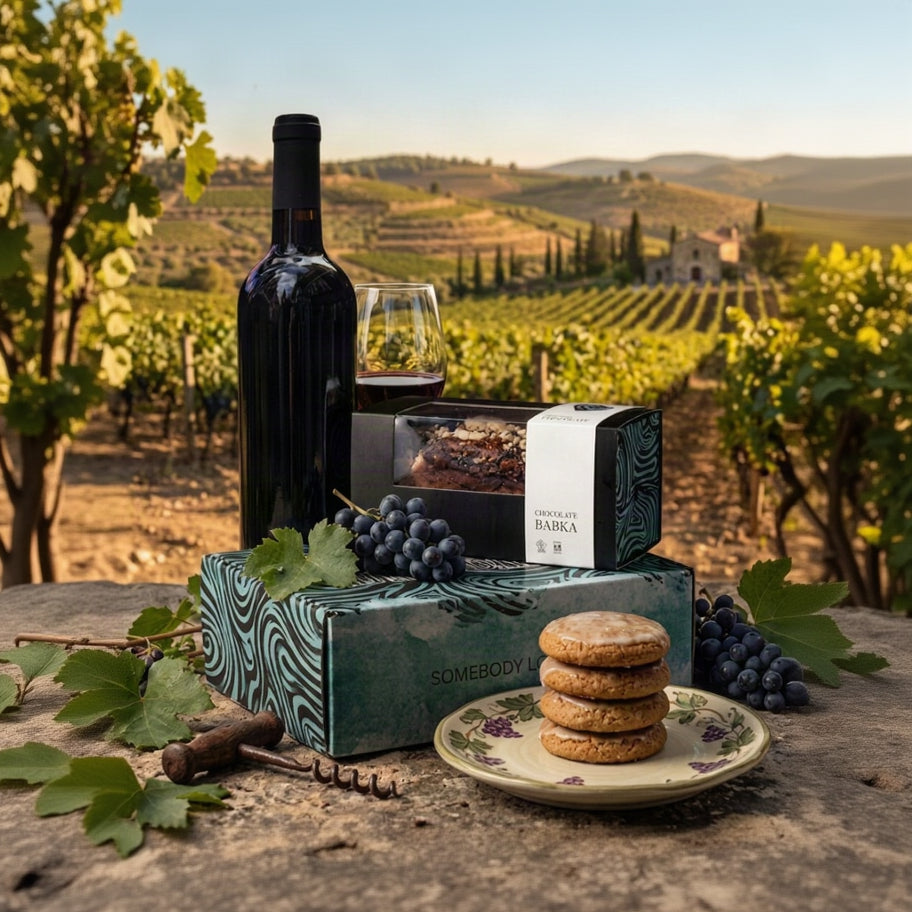 A curated gift set arranged outdoors on a stone surface with a vineyard landscape in the background. The set includes a dark wine bottle, a glass of red wine, a chocolate cake in a patterned box, a white box labeled 'CHOCOLATE FIORBA', a teal patterned gift box labeled 'SOMEBODY', grapes, cookies on a plate, and a corkscrew.