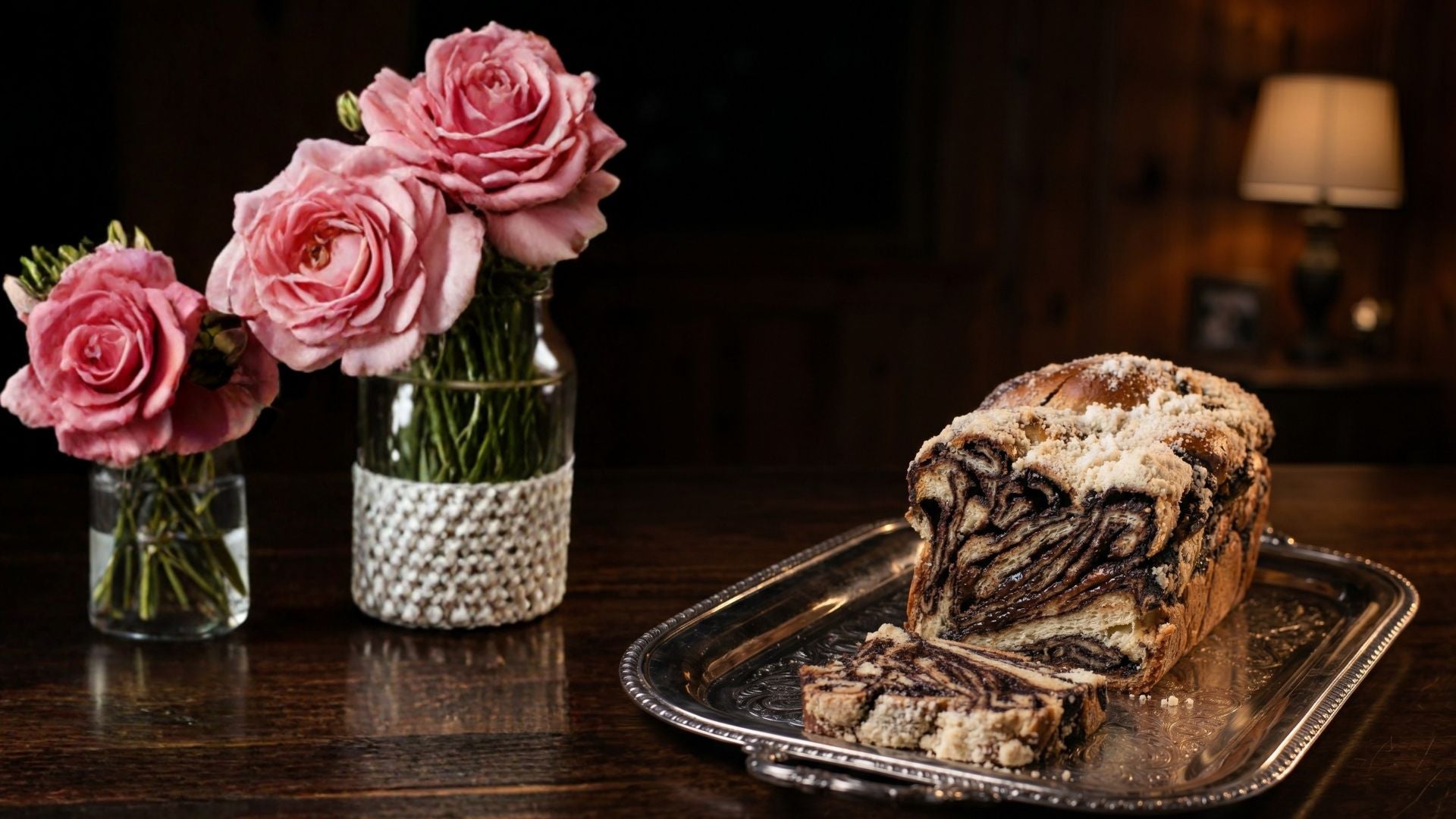 Slice of chocolate cake on a silver tray with pink roses in glass vases on a wooden table.
